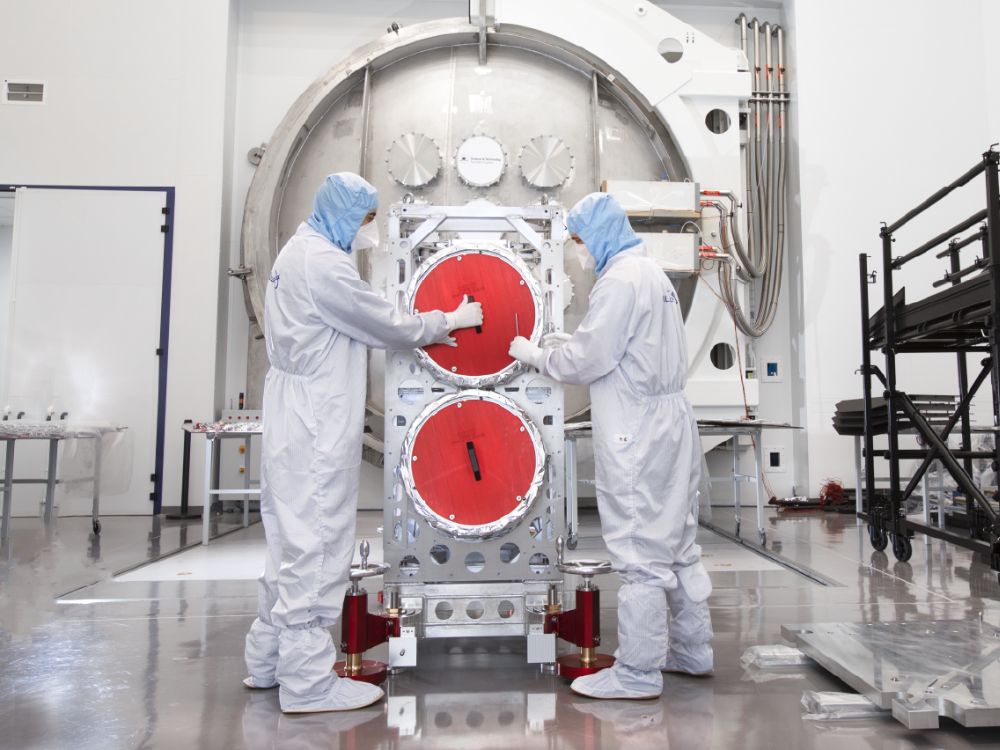 Two people wearing clean room suits standing in front of a thermal vacuum chamber door inspecting satellite components.